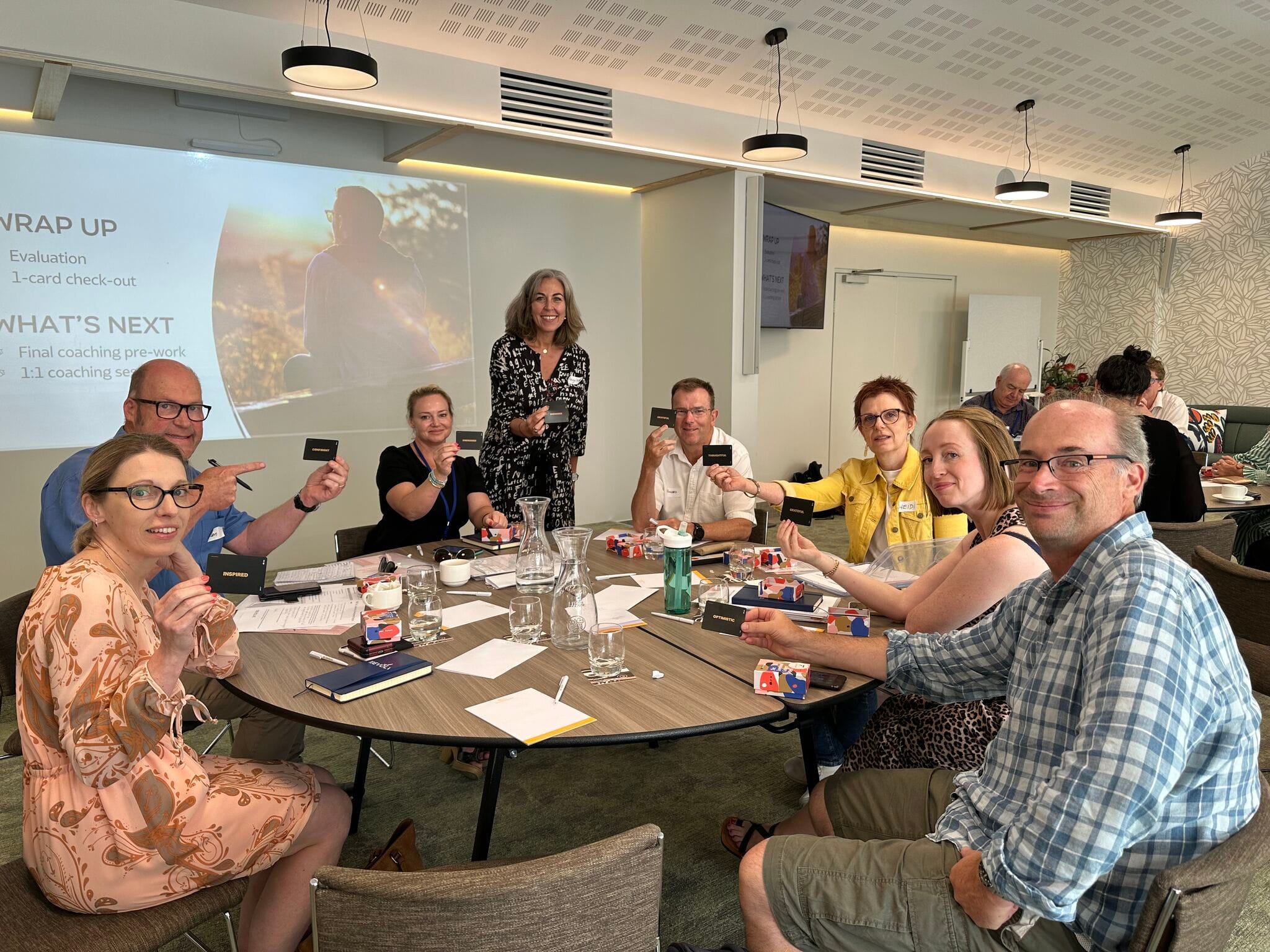 Angella Clarke-Jervoise A group of people sit around a table in a conference room, holding up cards, with a woman standing and a presentation projected on the screen behind them.