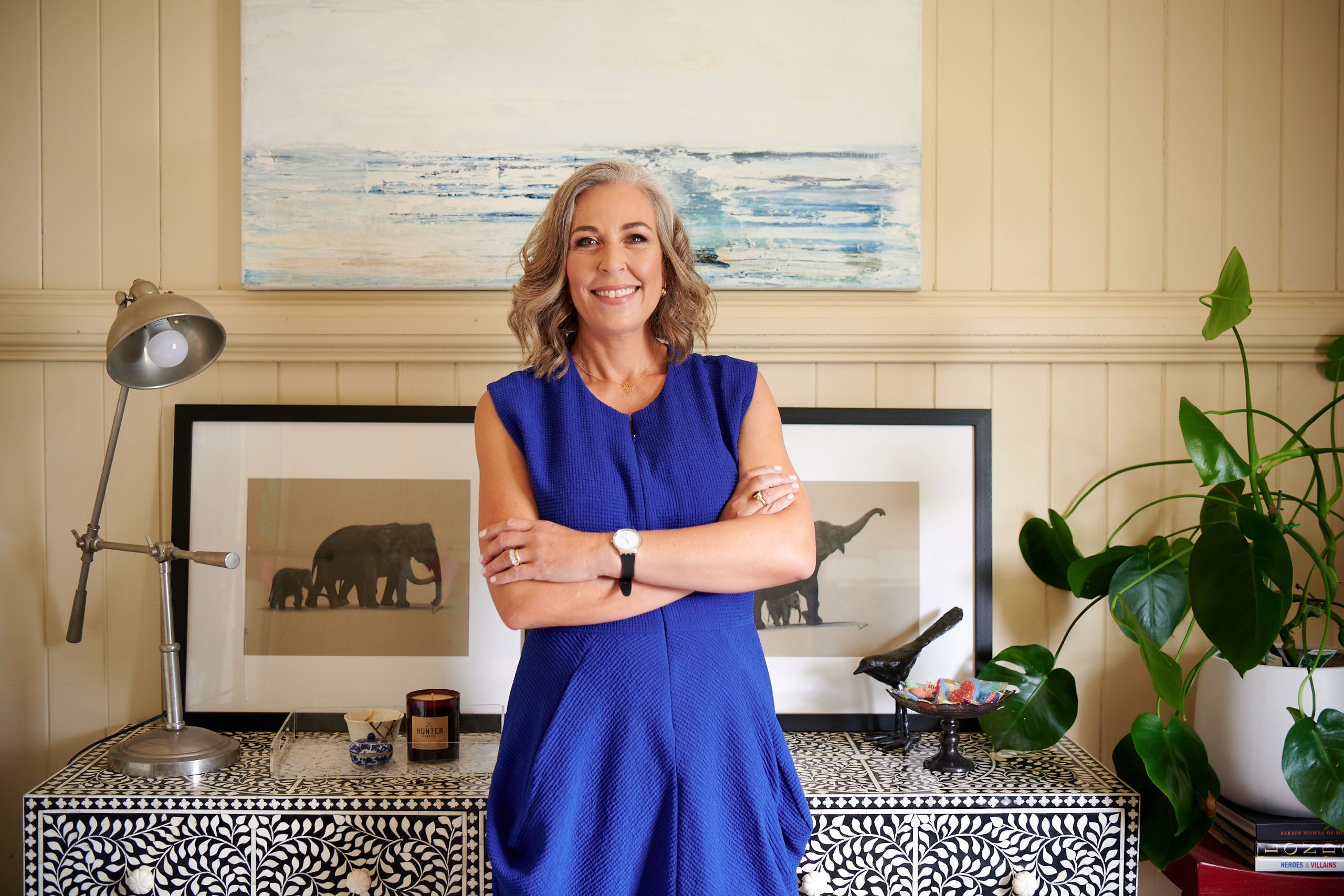 Angella Clarke-Jervoise A woman in a blue dress stands with arms crossed, smiling in a decorated room with framed art, a lamp, and a houseplant.