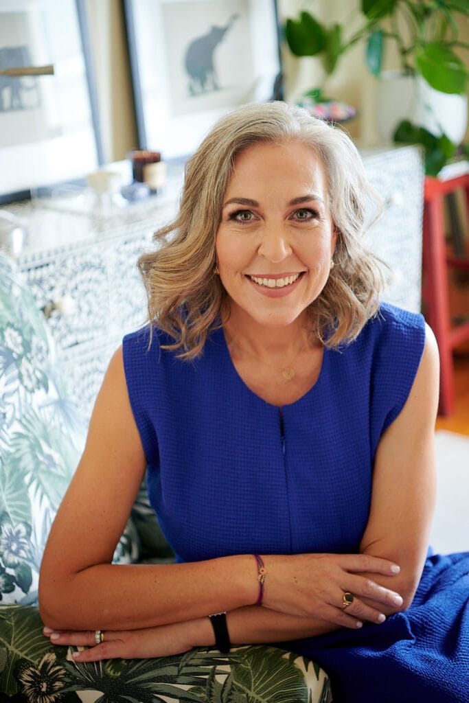 Angella Clarke-Jervoise A woman with shoulder-length gray hair wearing a blue dress sits on a patterned chair, smiling at the camera in a well-lit room with plants and framed artwork.