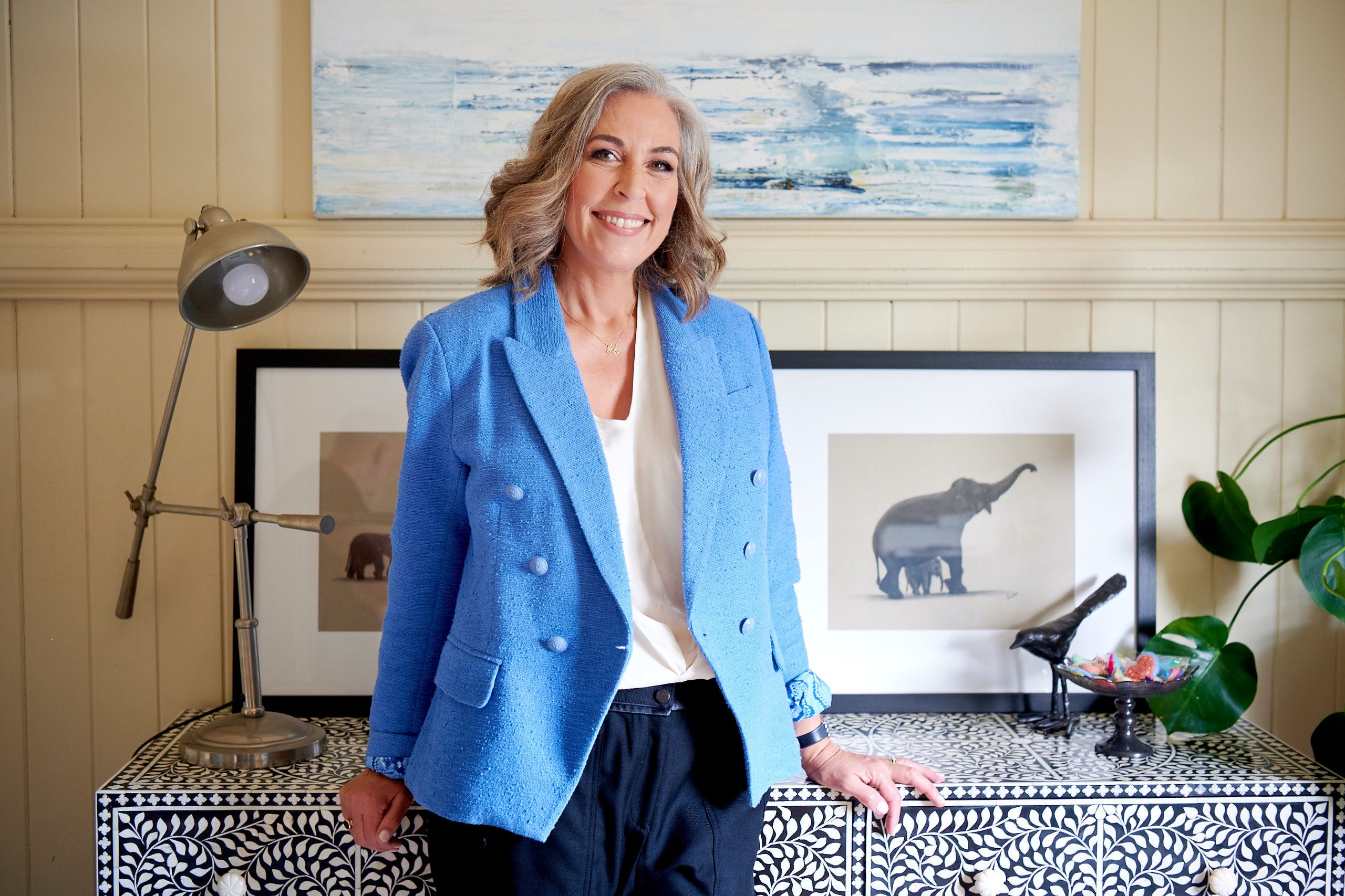 Angella Clarke-Jervoise A woman with gray hair in a blue blazer stands by a patterned cabinet, with framed elephant art and a lamp in the background.
