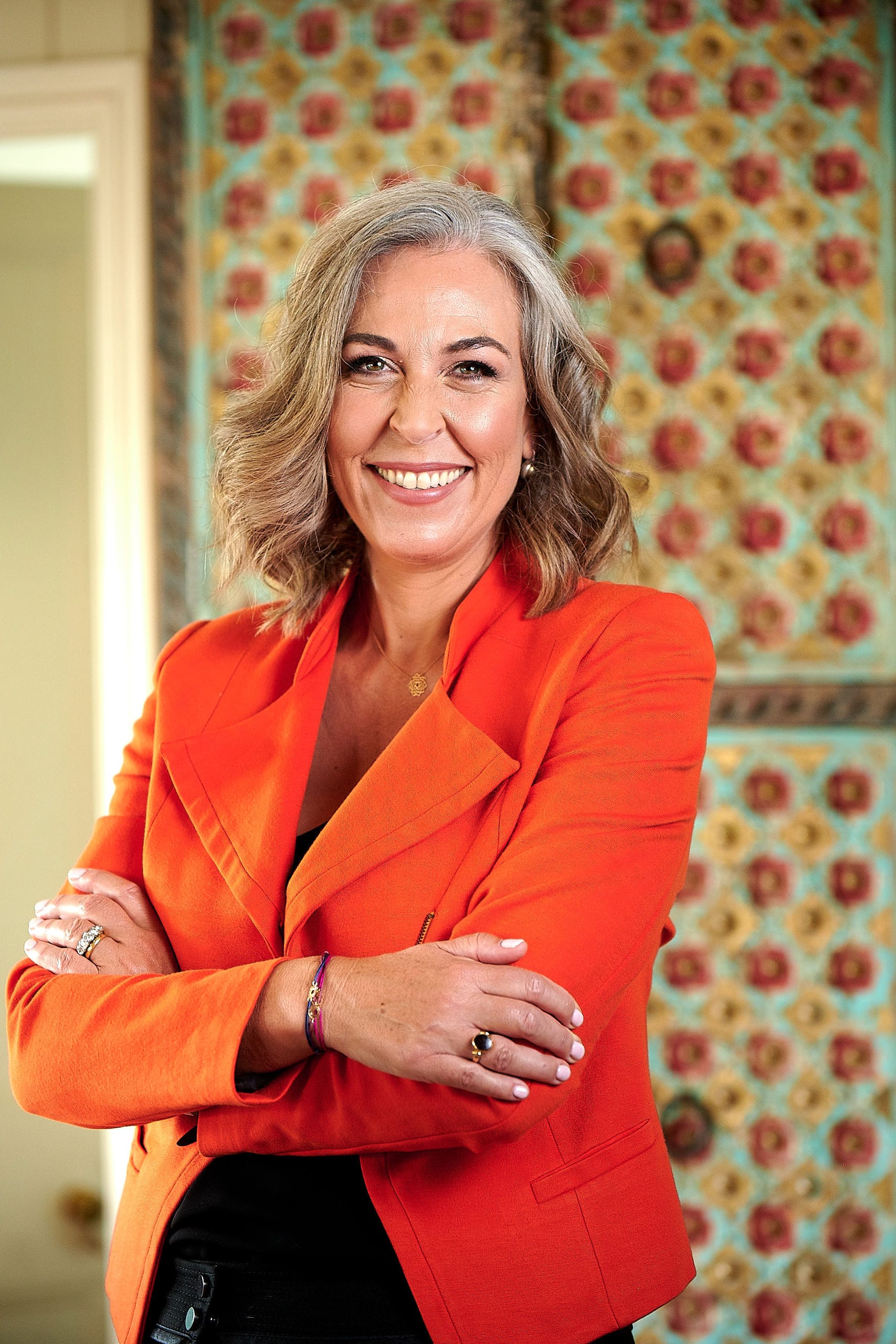 Angella Clarke-Jervoise A woman with wavy gray hair wearing an orange blazer stands smiling with arms crossed in front of a decorative, patterned background.