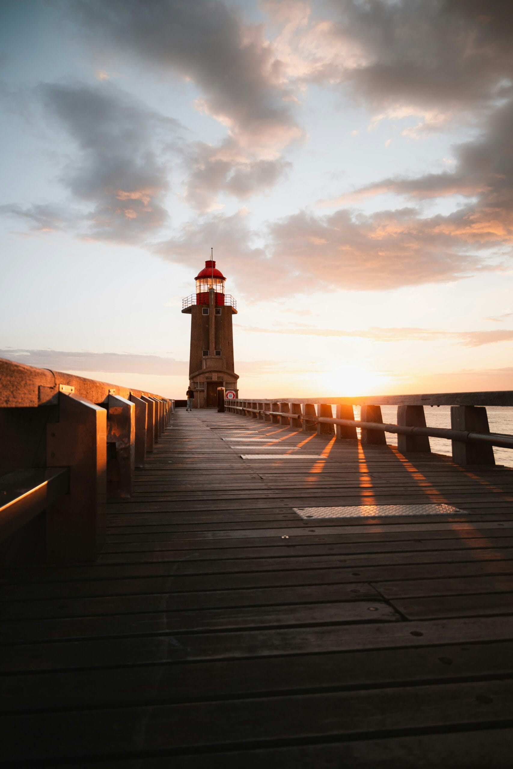 Angella Clarke-Jervoise A lighthouse stands at the end of a wooden pier at sunset, casting long shadows and reflecting light on the water.