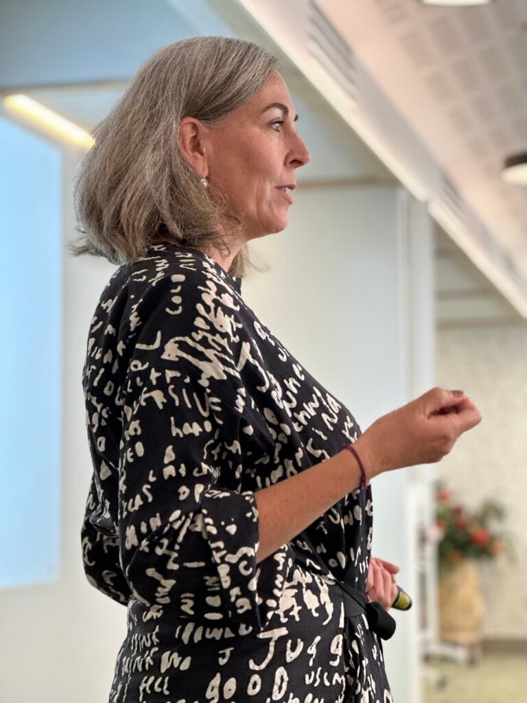 Angella Clarke-Jervoise A woman with gray hair wearing a patterned dress speaks while gesturing with her hand in a modern indoor setting.
