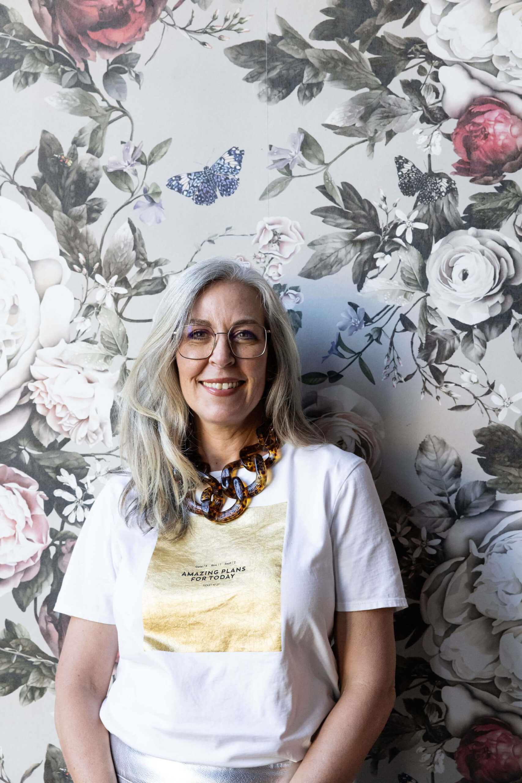 Angella Clarke-Jervoise A smiling woman with long gray hair and glasses stands in front of a floral wallpaper. She wears a white t-shirt with a gold square design and a chunky necklace. A blue butterfly image is on the wall behind her head.